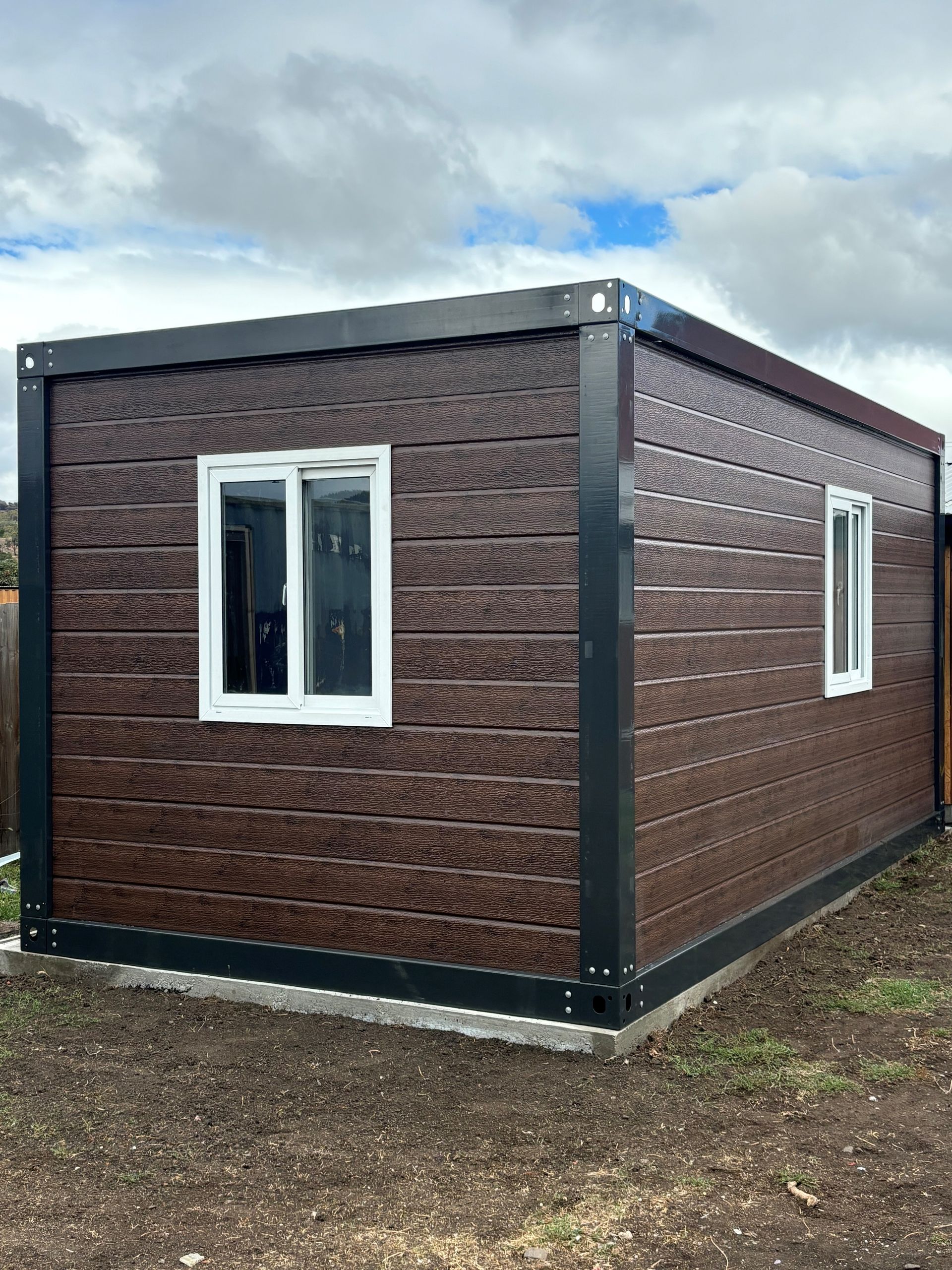 A brown house with two windows is sitting on top of a dirt field.