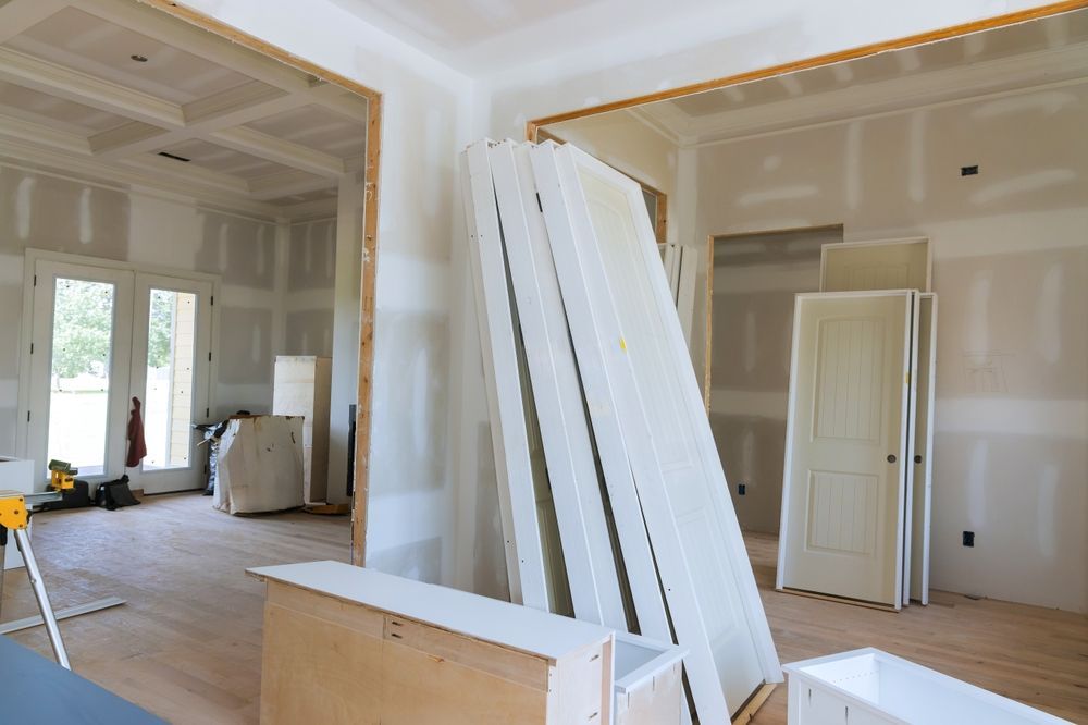 Interior of a house under renovation with drywall, stacked boards, and a doorway to a bright room