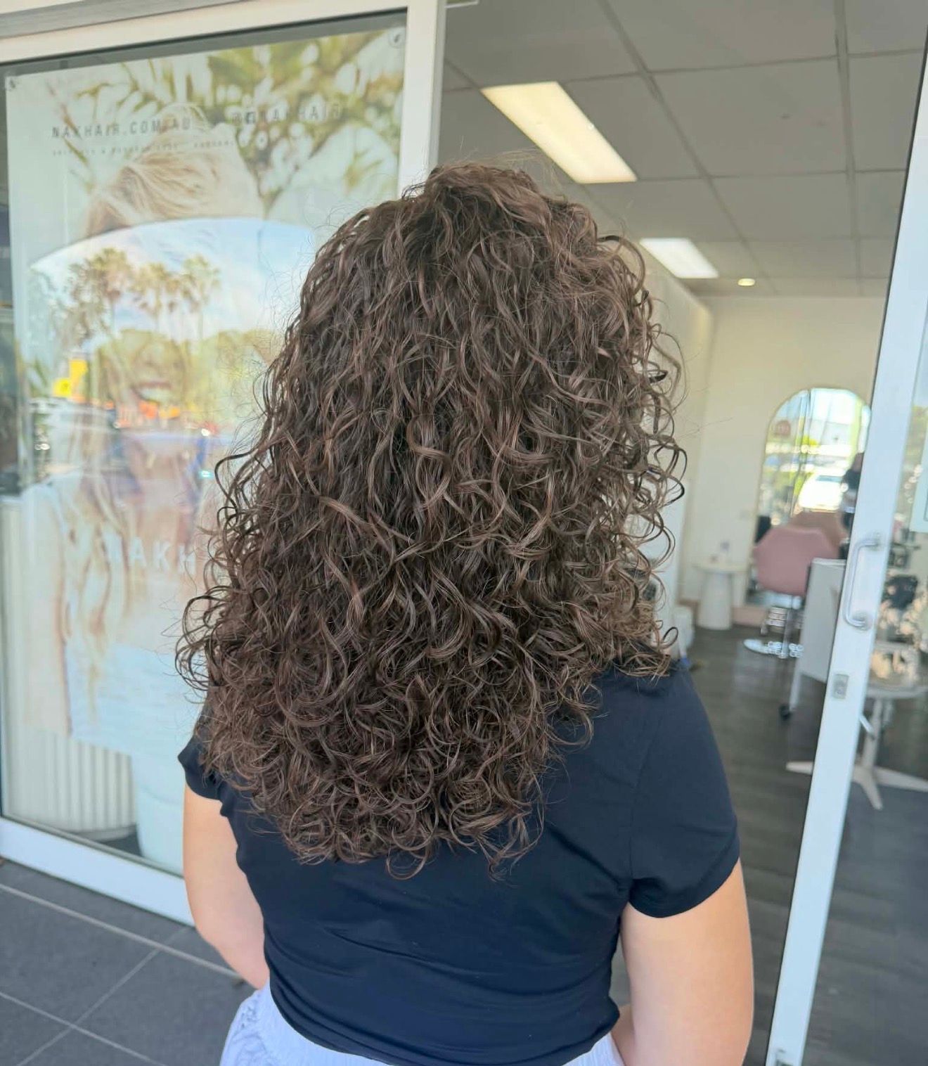 Person With Curly Brown Hair Standing Outside A Storefront, Seen From Behind — Oh So Blonde Hair Studio in Kawana, QLD