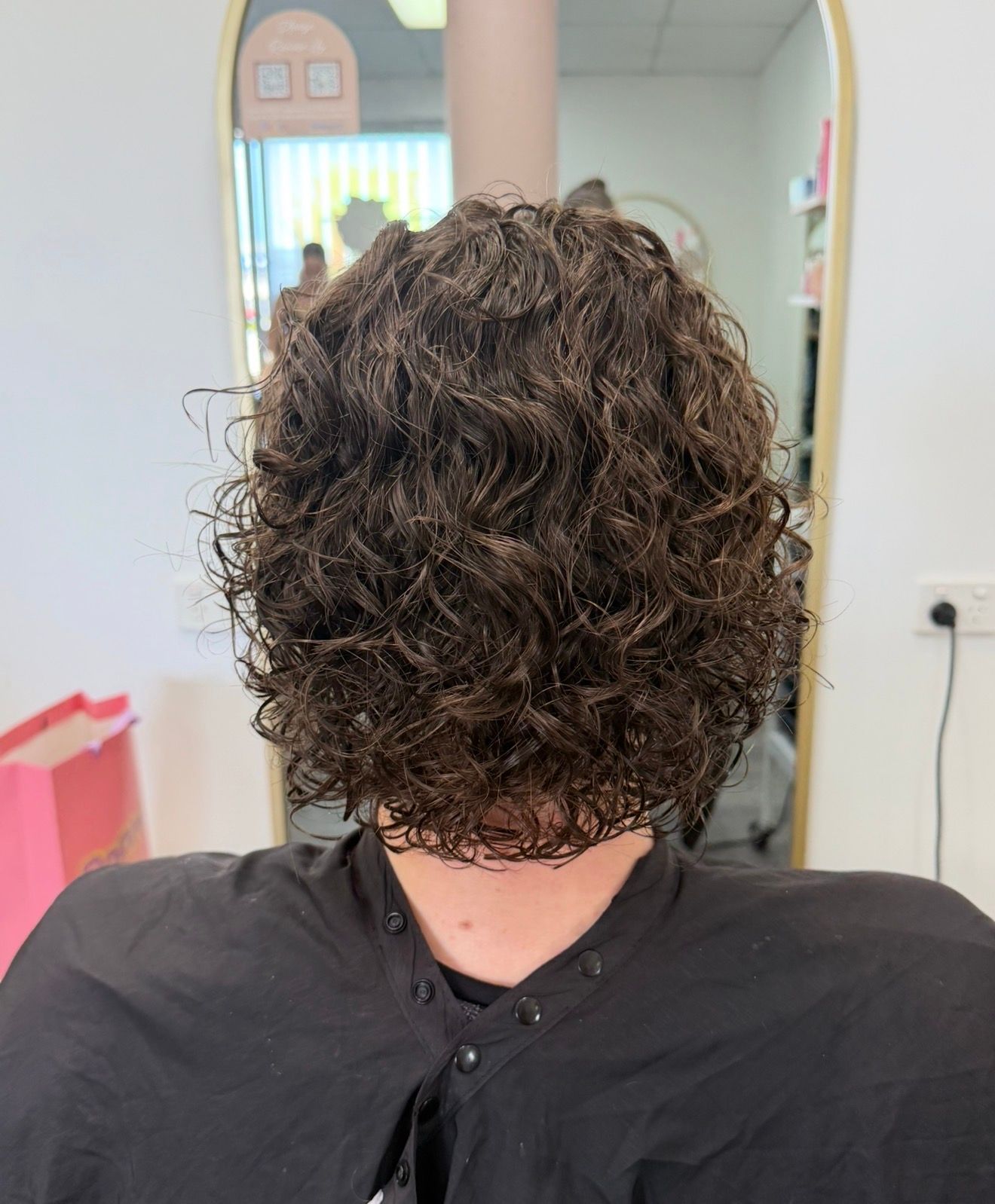 Person With Curly Brown Hair Shown From Behind In A Salon Chair, Wearing A Black Cape — Oh So Blonde Hair Studio in Buddina, QLD