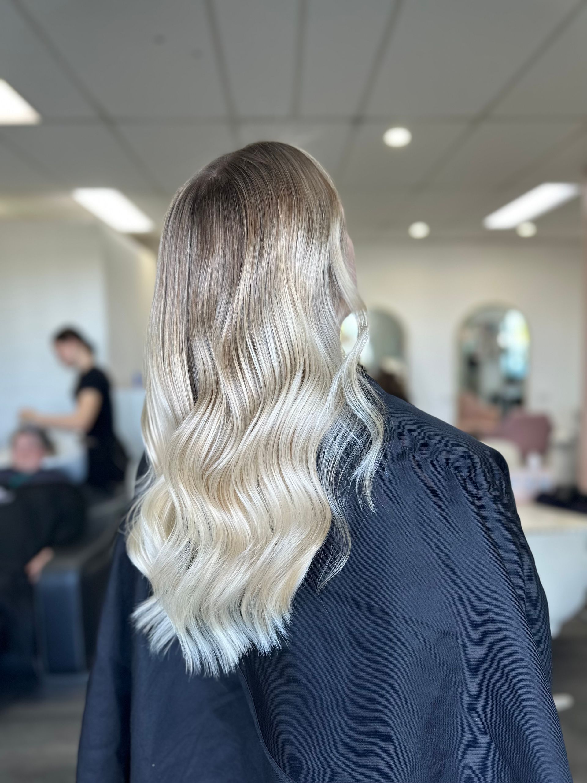 Blonde Wavy Hair Viewed From Behind In A Salon, With Blurred People And Mirrors In The Background — Oh So Blonde Hair Studio in Kawana, QLD