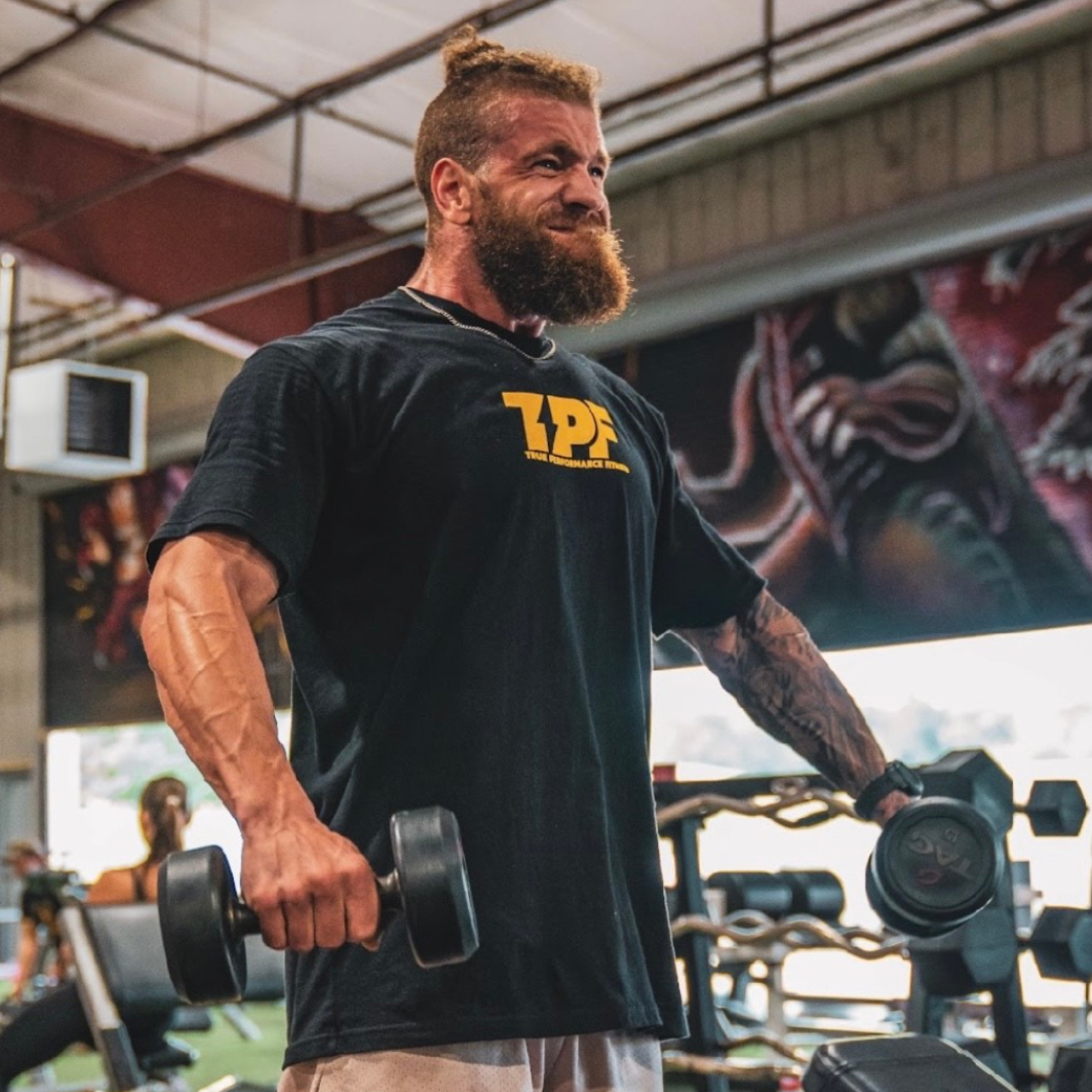 Man with beard lifting dumbbells in a gym. Black t-shirt with logo.