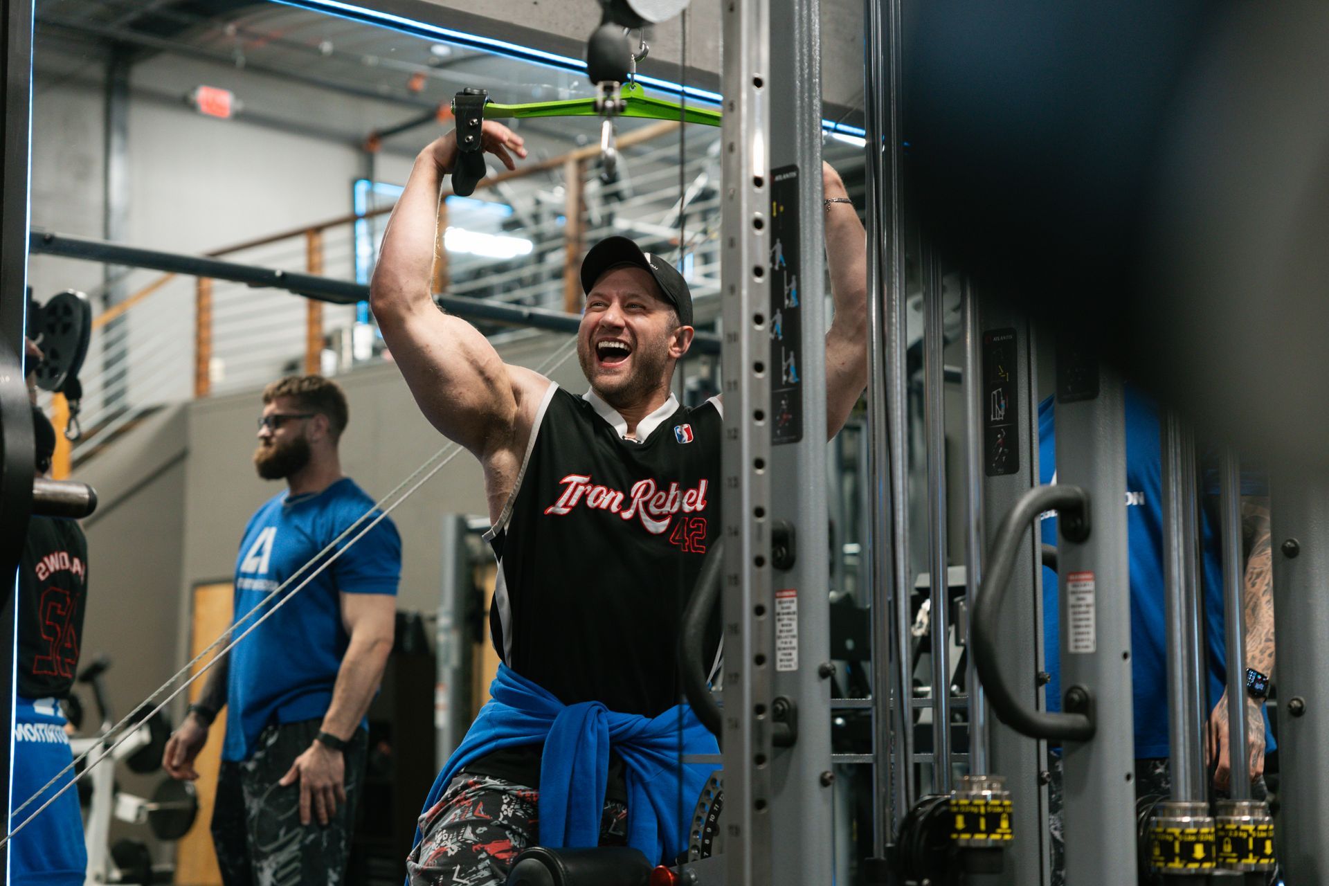 Man in gym, arms raised, celebrating workout. Another man in the background, also in the gym.