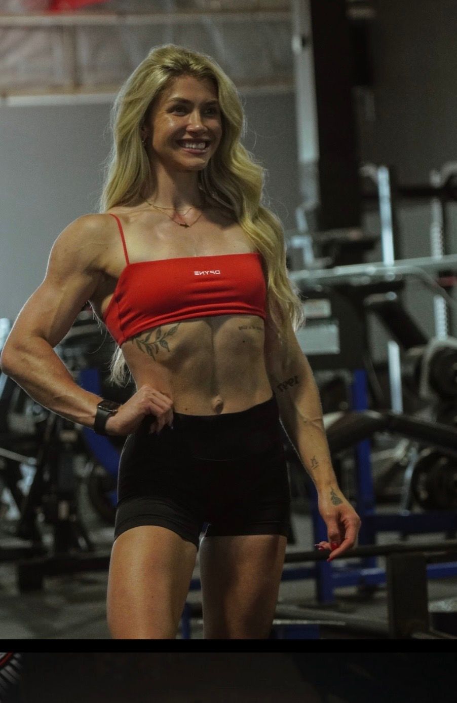 A smiling individual in a red top and black shorts stands in a gym, showing muscular definition.