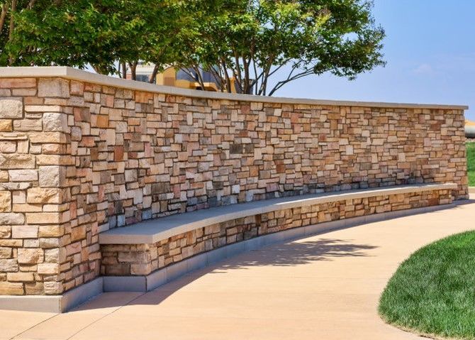Curved stone bench built into a tan stone wall along a sidewalk beside a grassy area