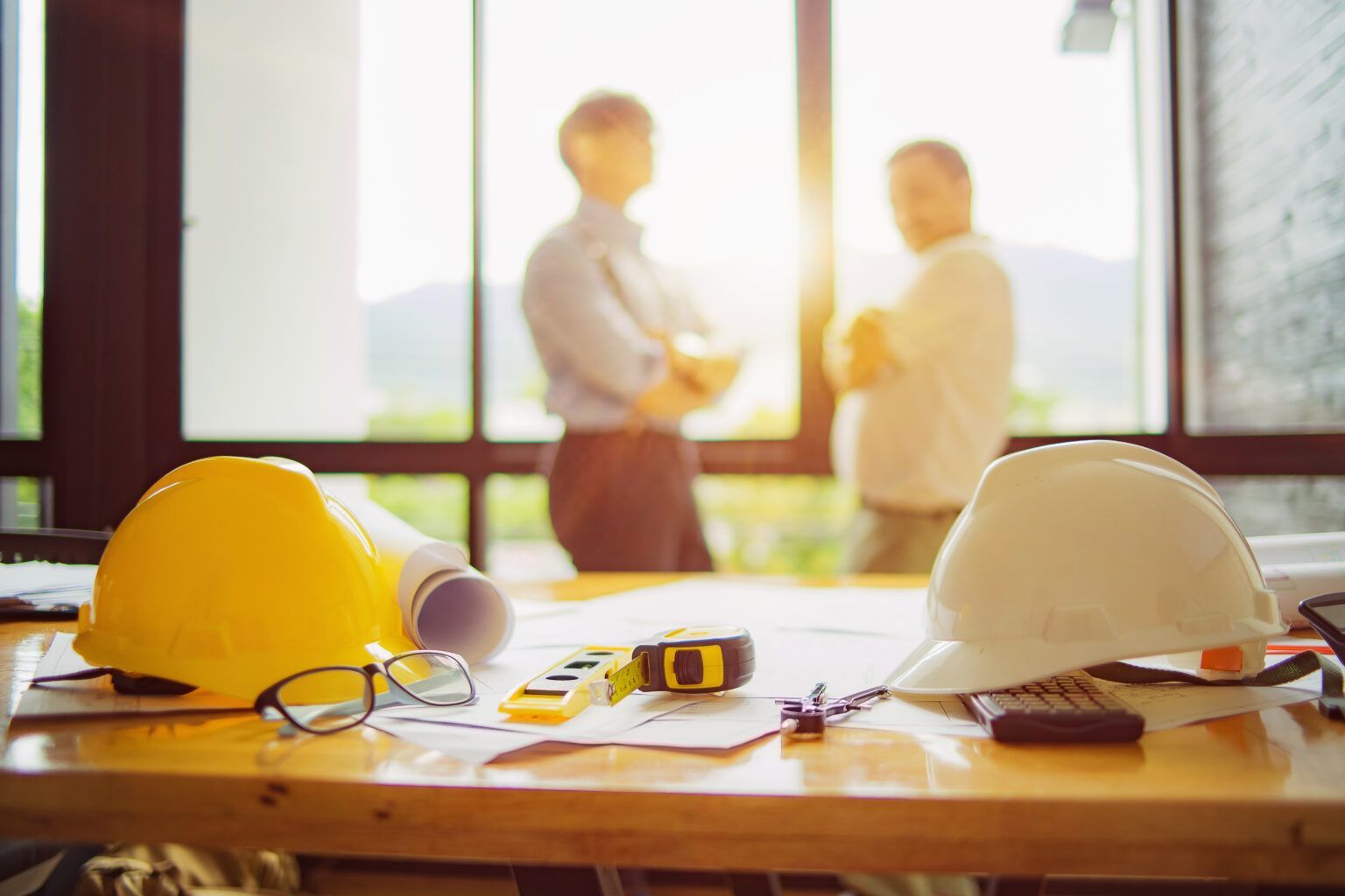 Construction workplace with two blurred figures near a window. Yellow and white hard hats, blueprints, and tools on a table.
