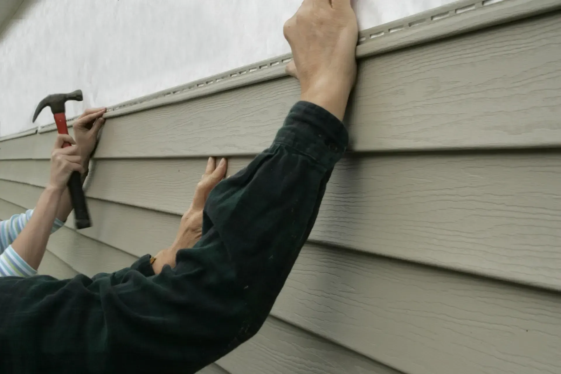 Person installing light brown siding on a building, using a hammer to nail it in.