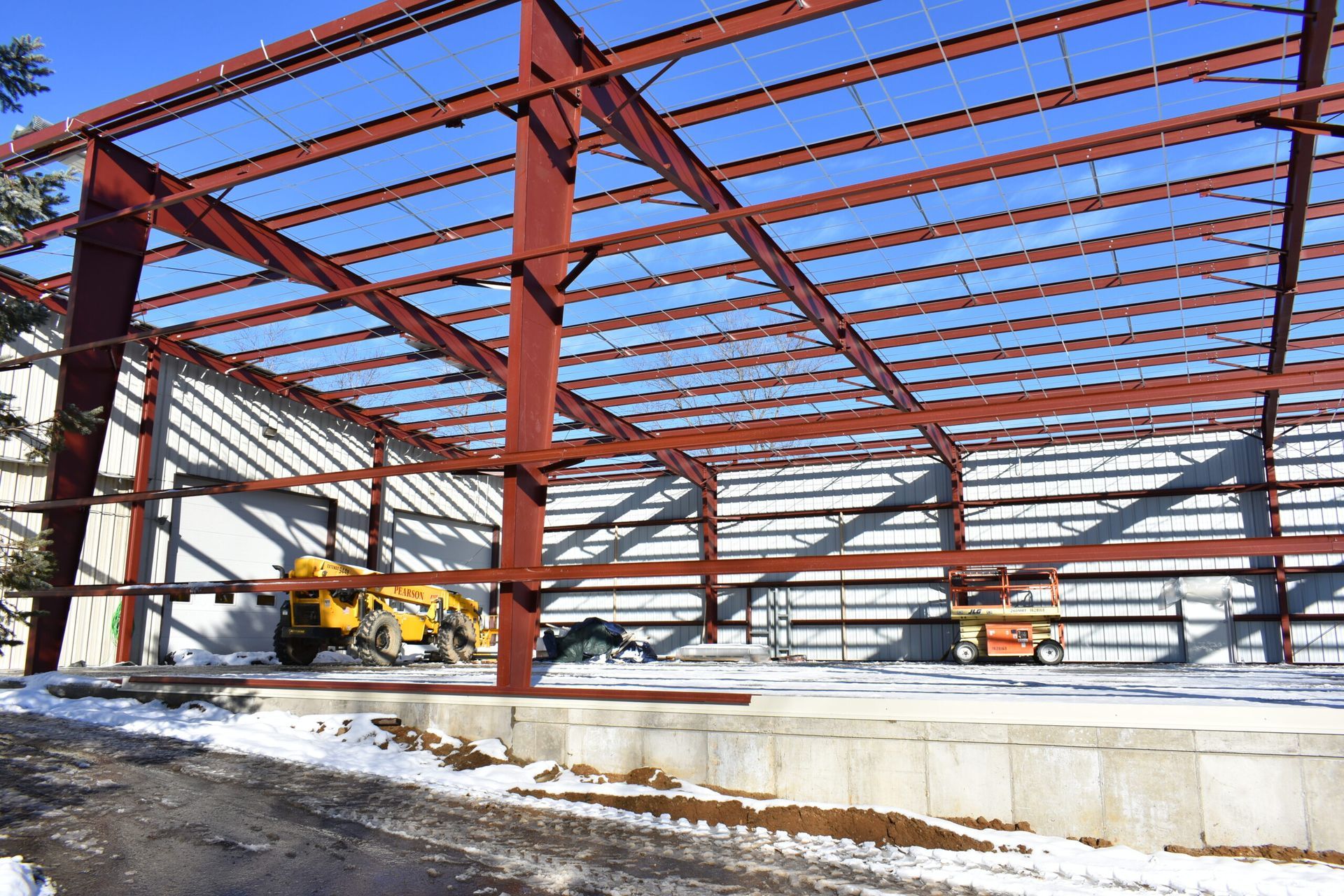 Steel frame of a building under construction, red-brown beams against a blue sky, snow on the ground.