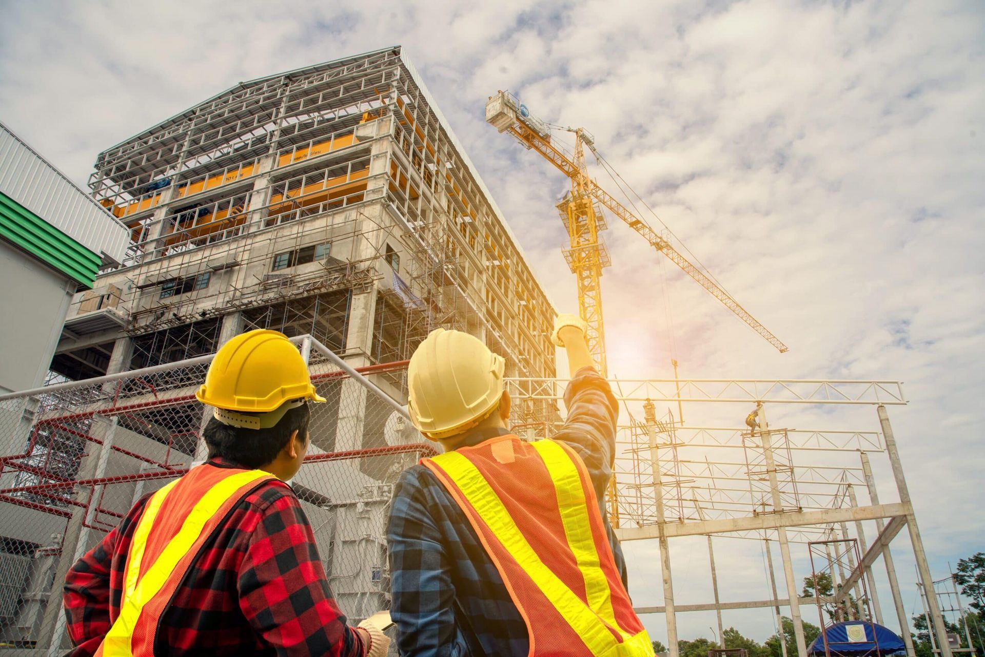 Two construction workers in yellow hard hats and vests looking up at a building under construction.