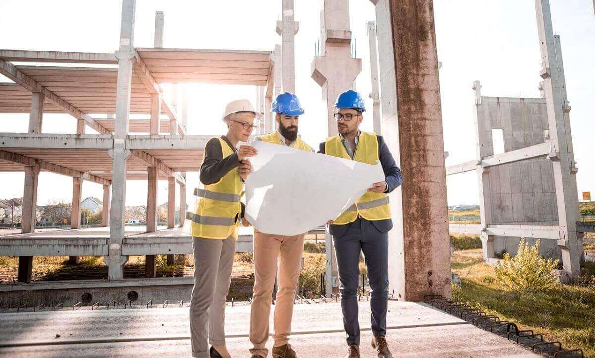 Three construction workers in hard hats examine blueprints at a building site.