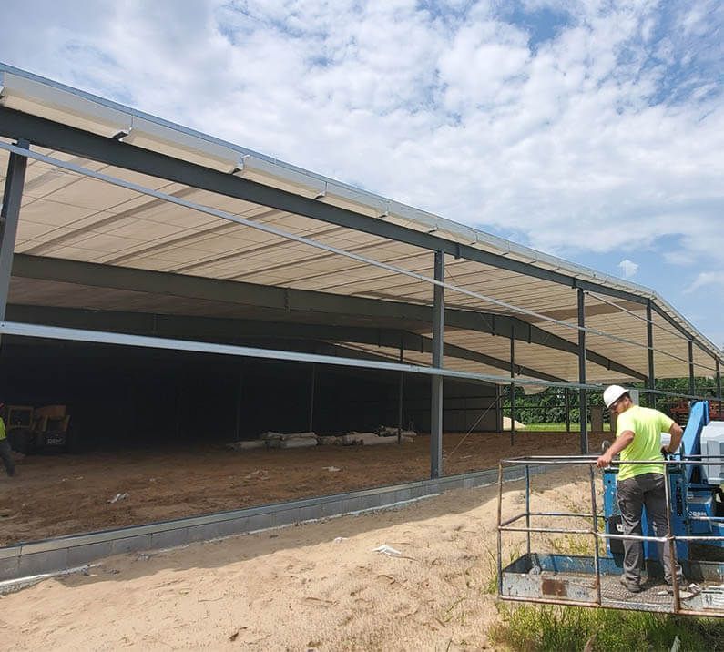 Construction of a metal building with workers, including one in a lift. Brown dirt and blue sky.