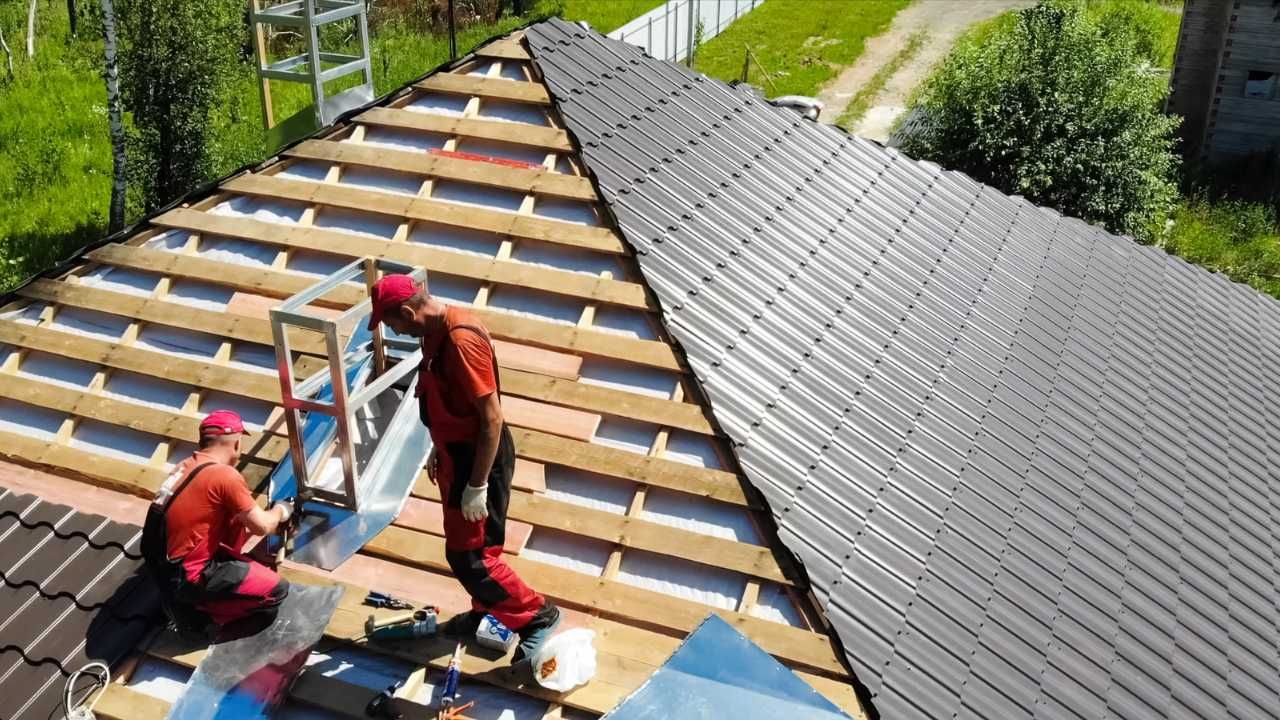 Two roofers in red overalls installing metal roof on a house.