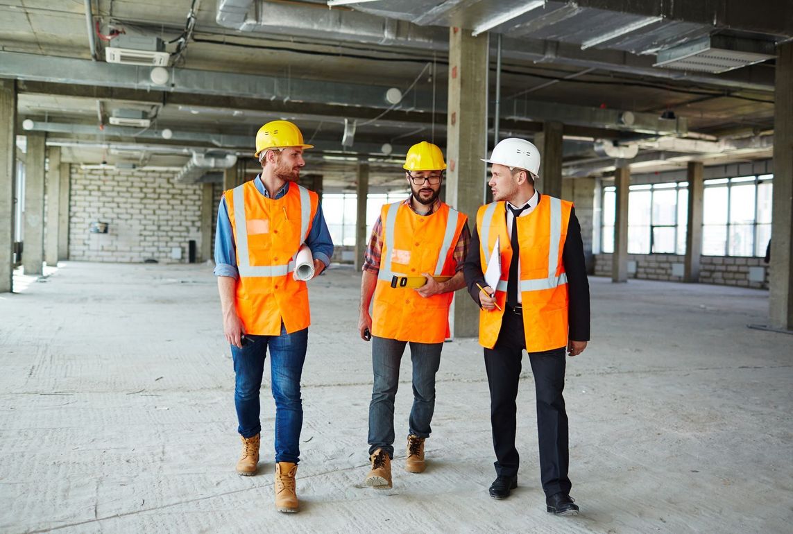 Three construction workers in hard hats and vests walking inside a building under construction.