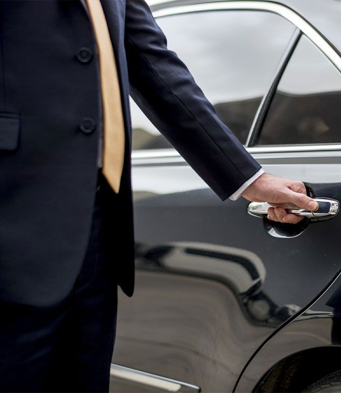 A person in a dark suit and gold tie reaches to open the door of a black car.