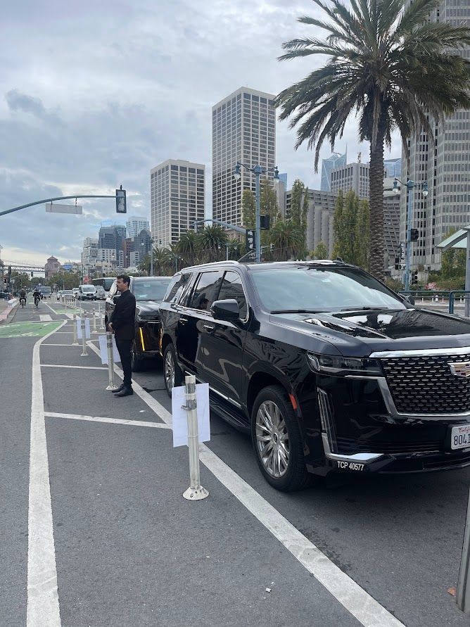 A black Cadillac SUV is parked in a bike lane on a city street, partially obstructing the path behind traffic bollards.