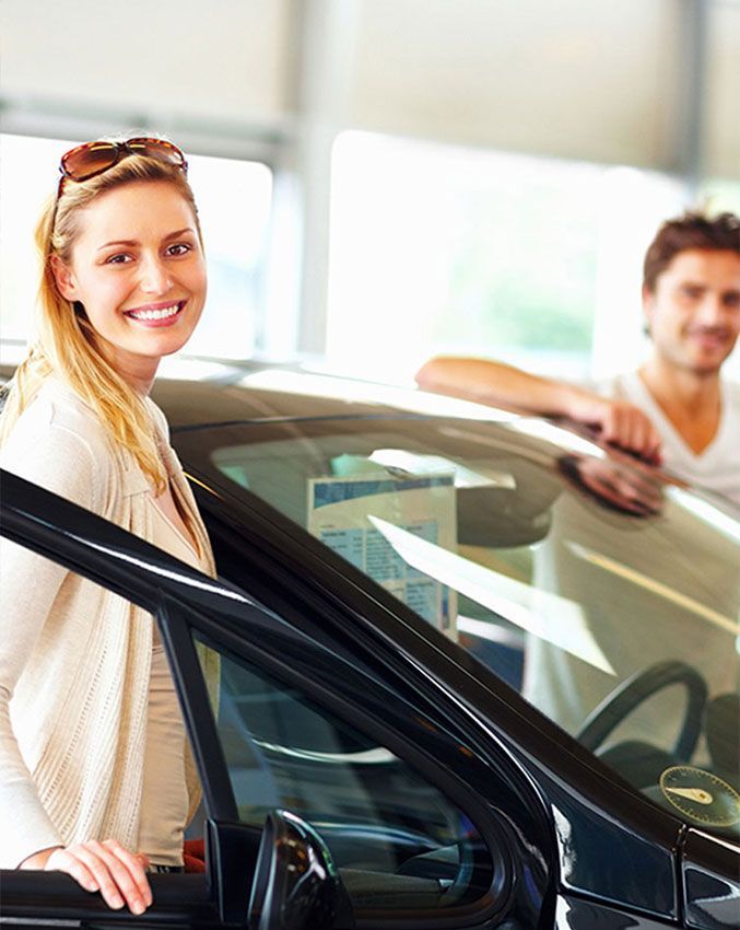 A smiling person stands by an open car door in a showroom, while another person leans against the vehicle in the background.
