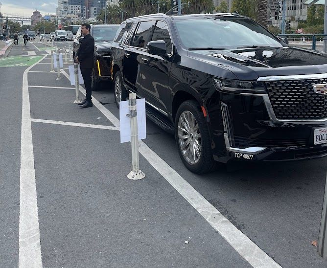 A black Cadillac SUV is parked in a bike lane on a city street, partially obstructing the path behind traffic bollards.