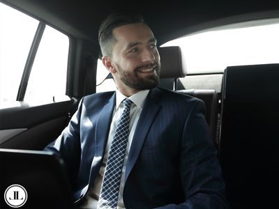 A smiling professional in a suit and tie sitting in the back seat of a car while working on a laptop.