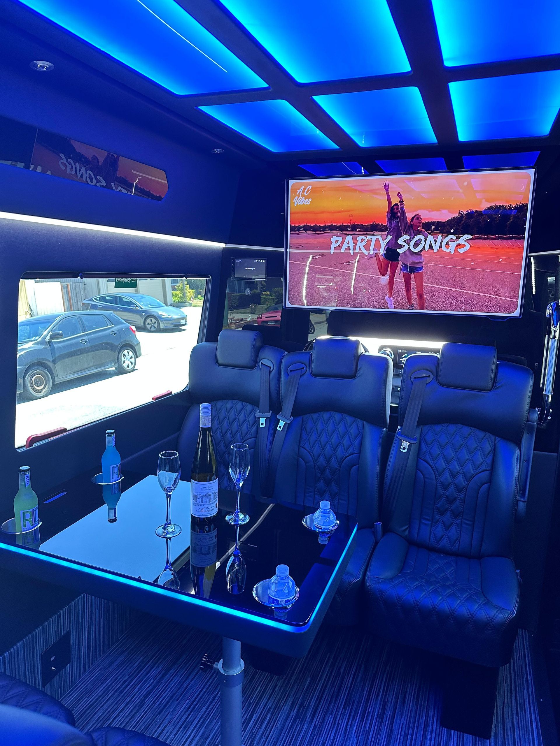 Interior of a party bus featuring blue ambient lighting, a dining table with glasses, and a television screen.