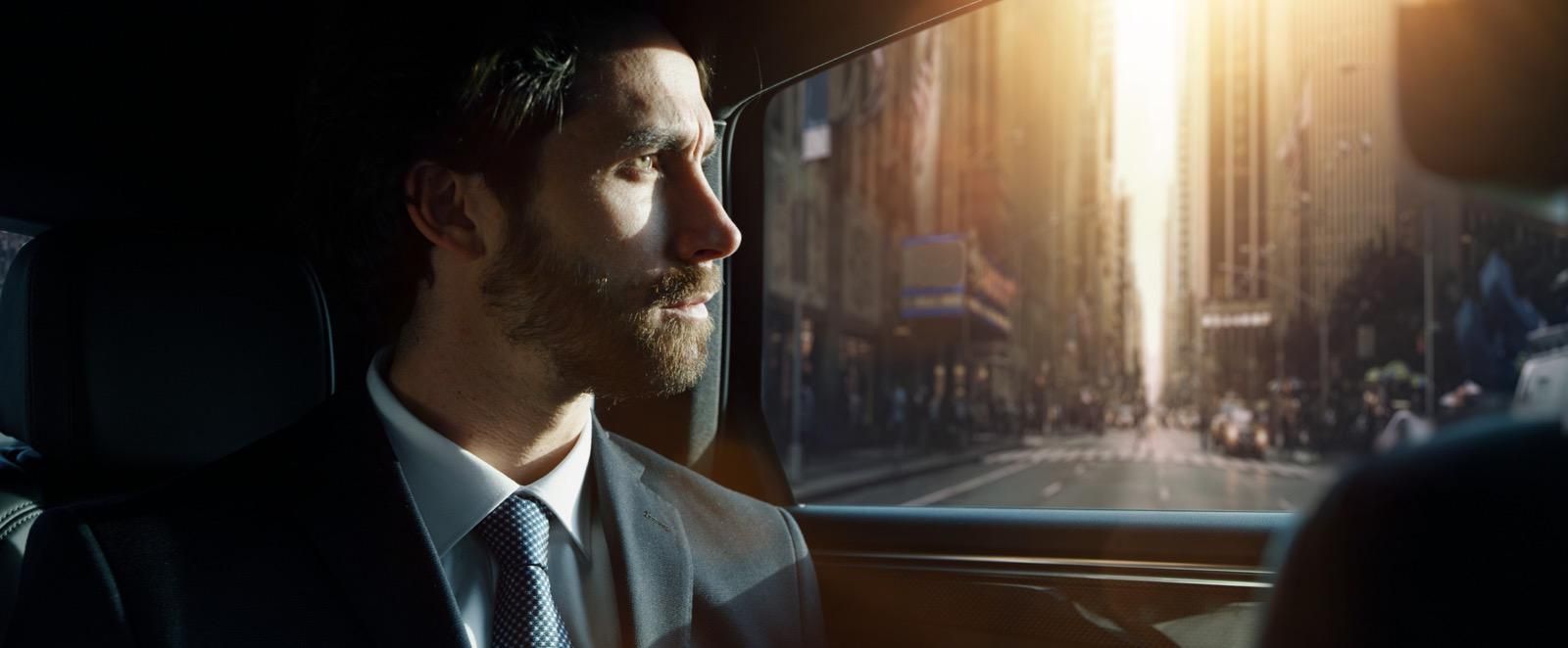 A man in a suit sits in the back of a car, looking out the window at a bright, sunny city street.