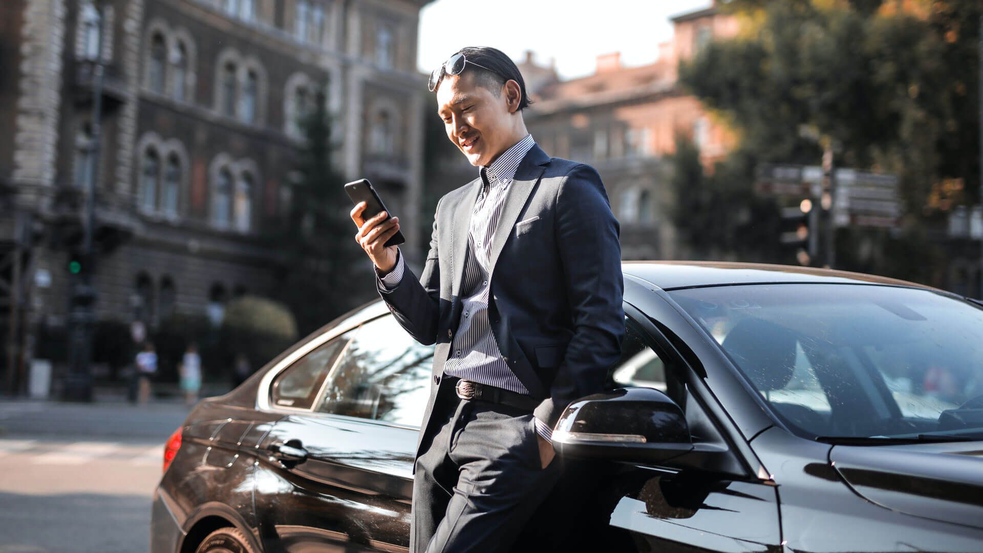 A person in a suit leans against a black car on a city street, smiling while looking at a smartphone.