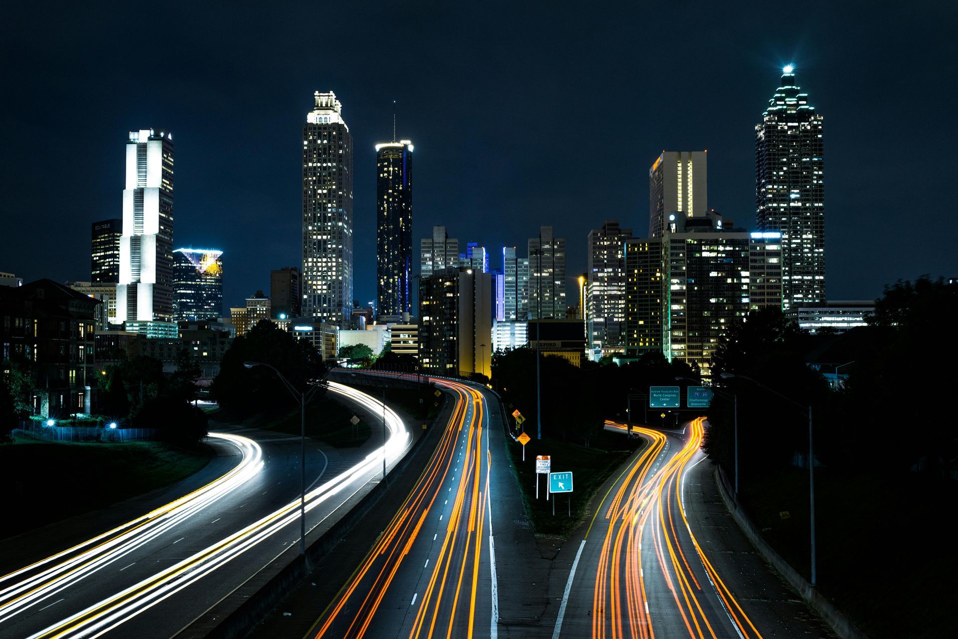 A night view of the Atlanta skyline with light trails from cars moving along a multi-lane highway in the foreground.