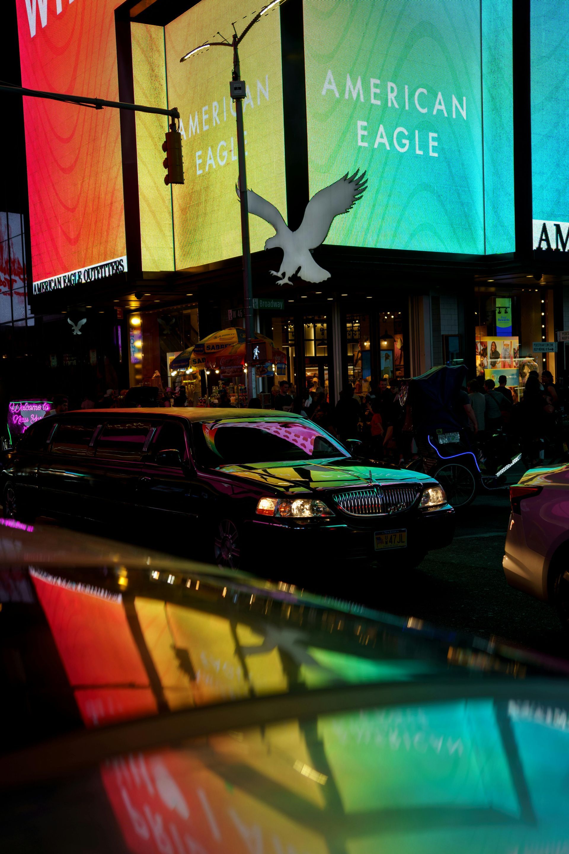 A black limousine drives past a brightly lit American Eagle store in a city at night, with reflections on the hood.