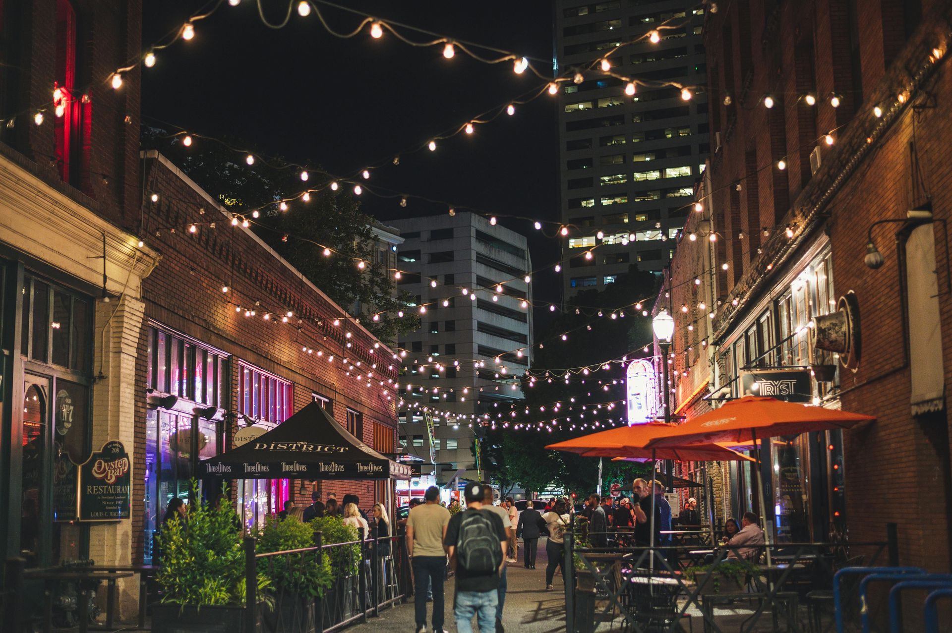 People gather in an urban alley at night, illuminated by decorative string lights and orange patio umbrellas.