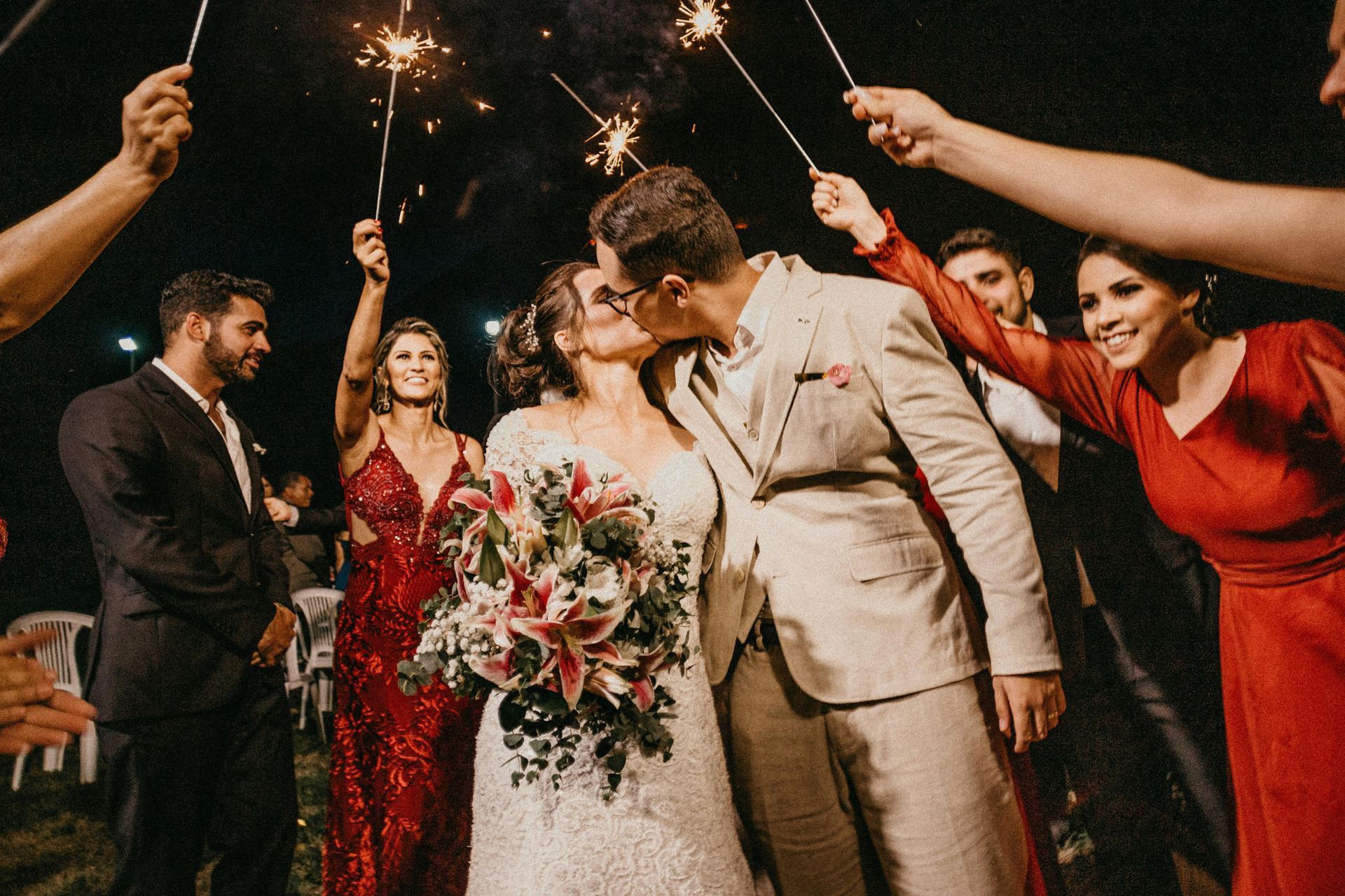 A wedding couple kissing under sparkling fireworks held by friends at an outdoor night reception.