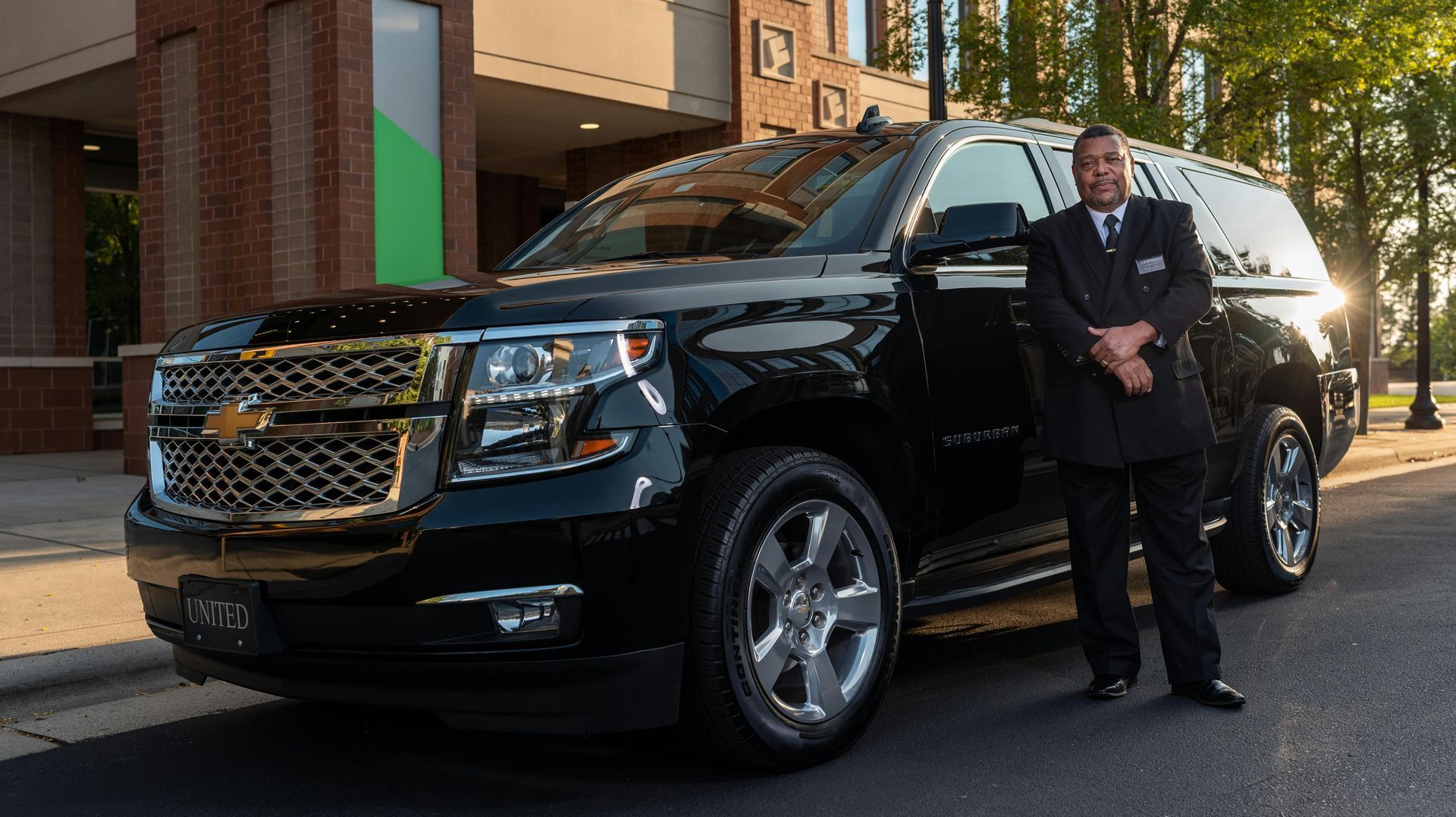 A driver in a suit stands beside a black SUV parked on a street outside a building.