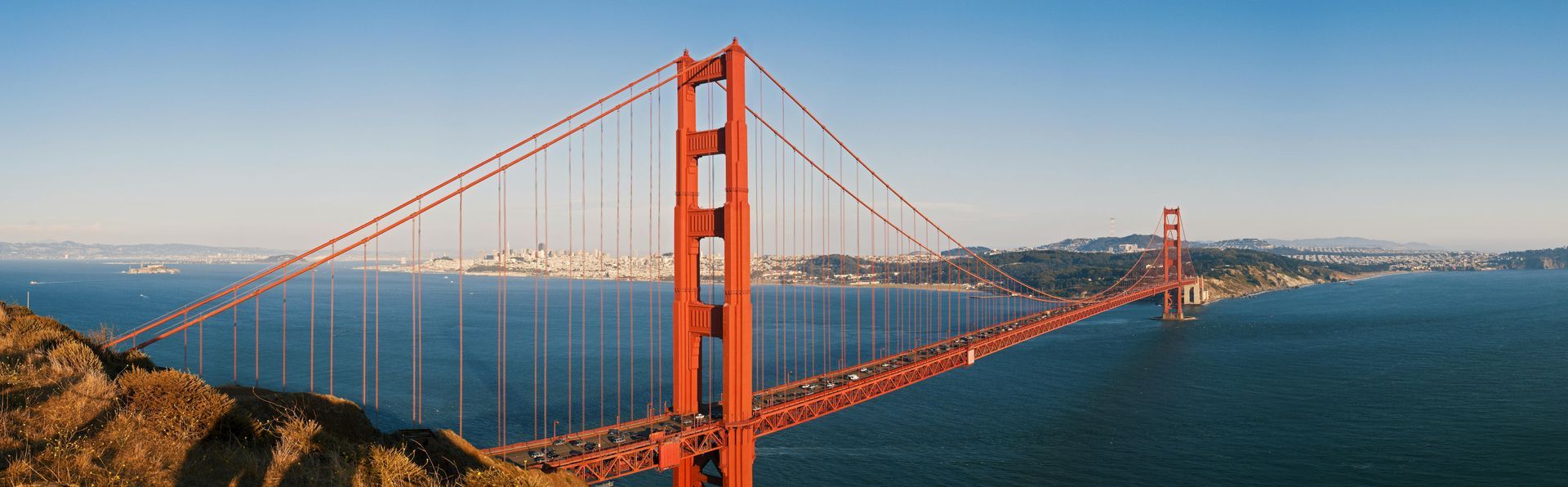 Wide-angle view of the bright orange Golden Gate Bridge spanning a blue bay under a clear sky.