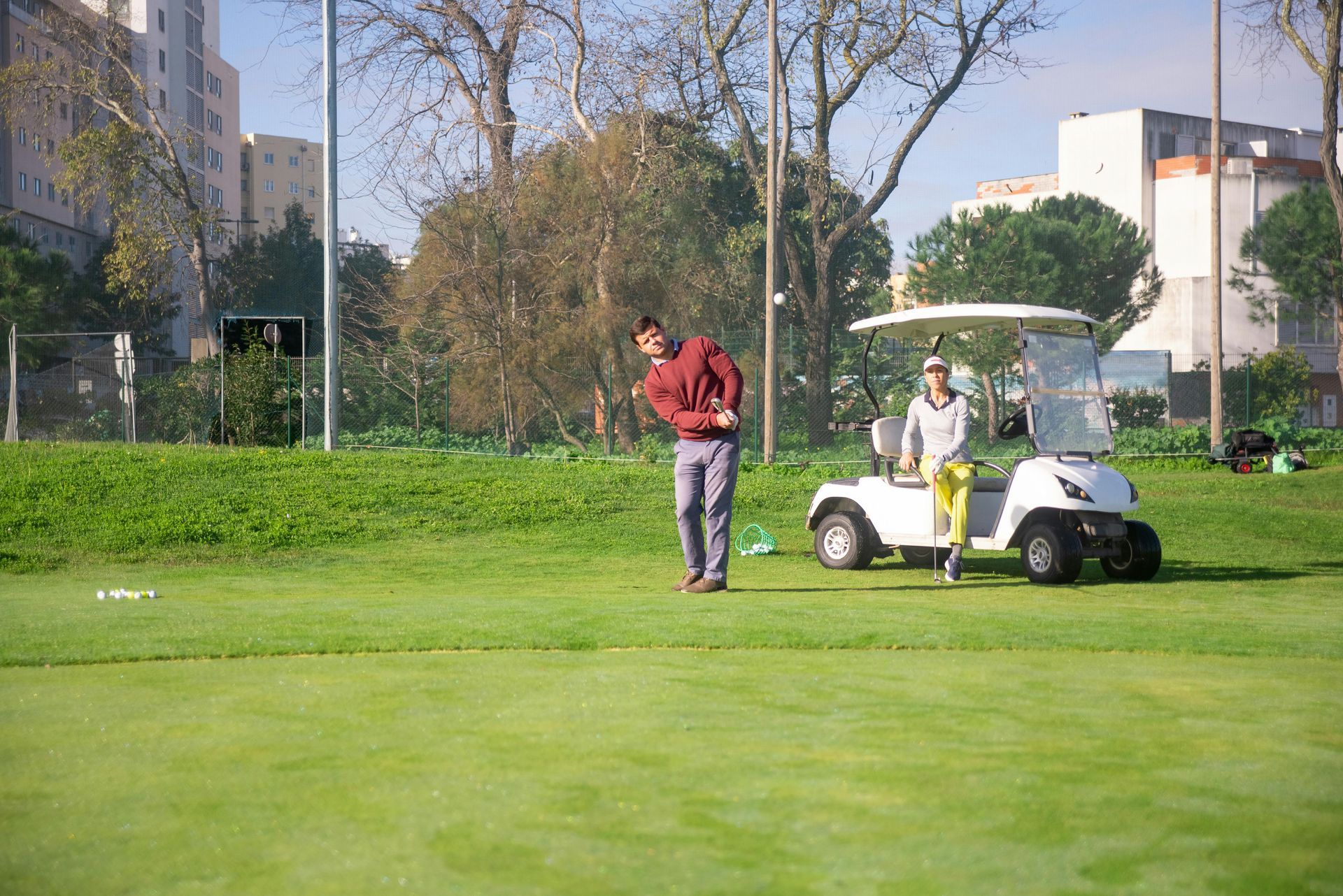 A golfer in a maroon sweater prepares to hit a ball on a green, while a person sits in a parked white golf cart nearby.