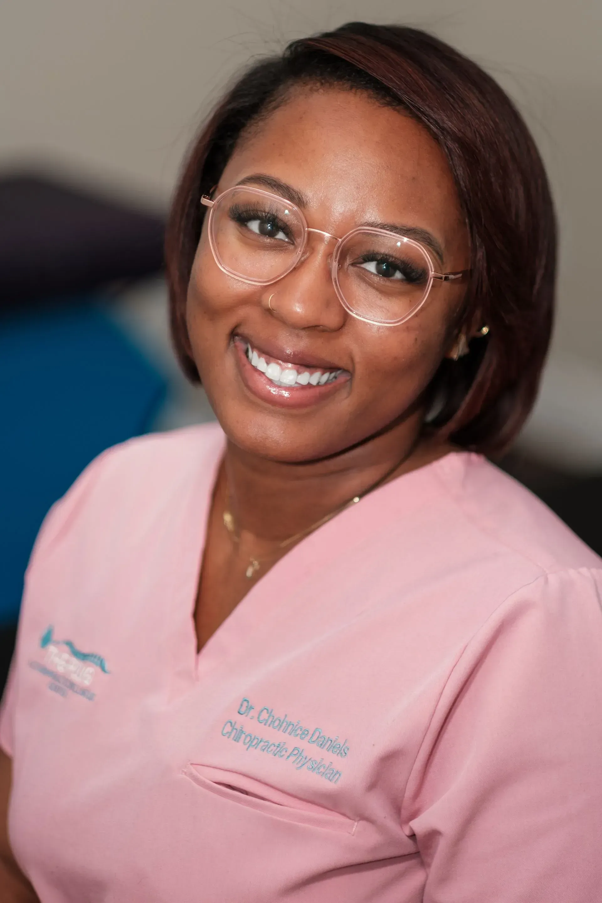 Woman with glasses and a white blouse smiles in front of a brick wall. She is wearing a smartwatch and has her arms crossed.