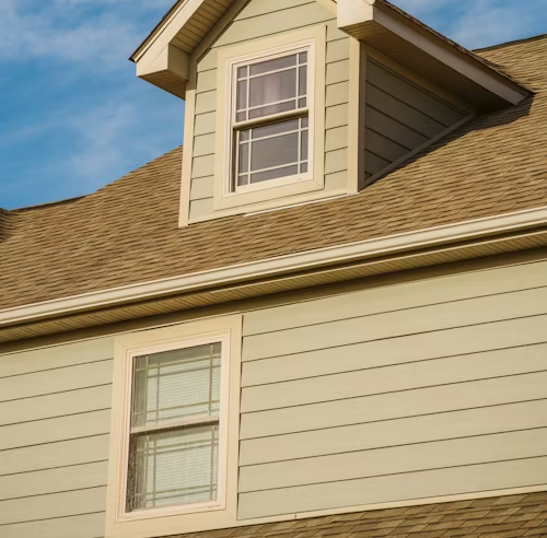 A close-up of a house exterior with light green horizontal siding, tan roof shingles, and two rectangular windows.
