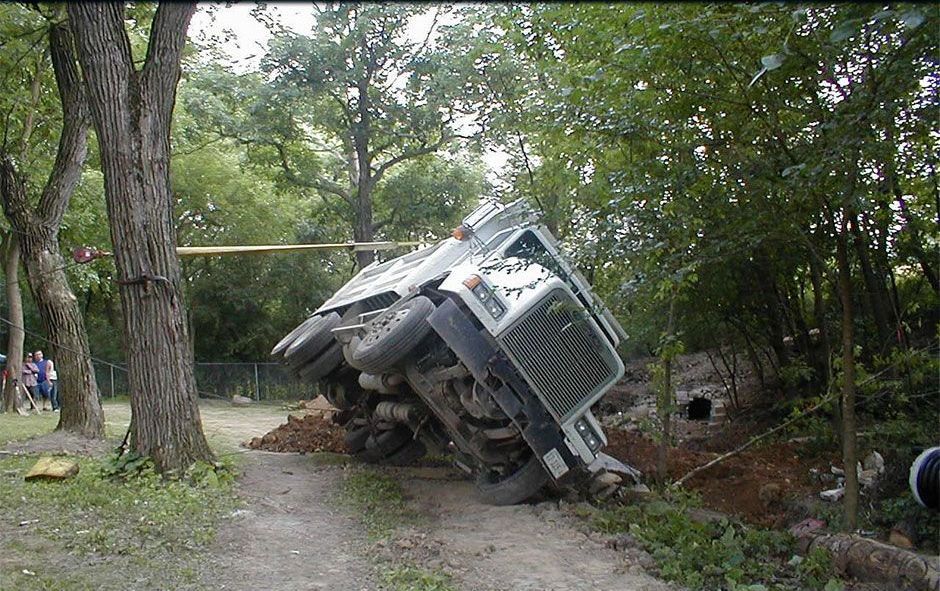 An overturned military-style vehicle on a dirt road near trees and vegetation.