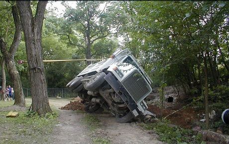 An overturned military-style vehicle on a dirt road near trees and vegetation.