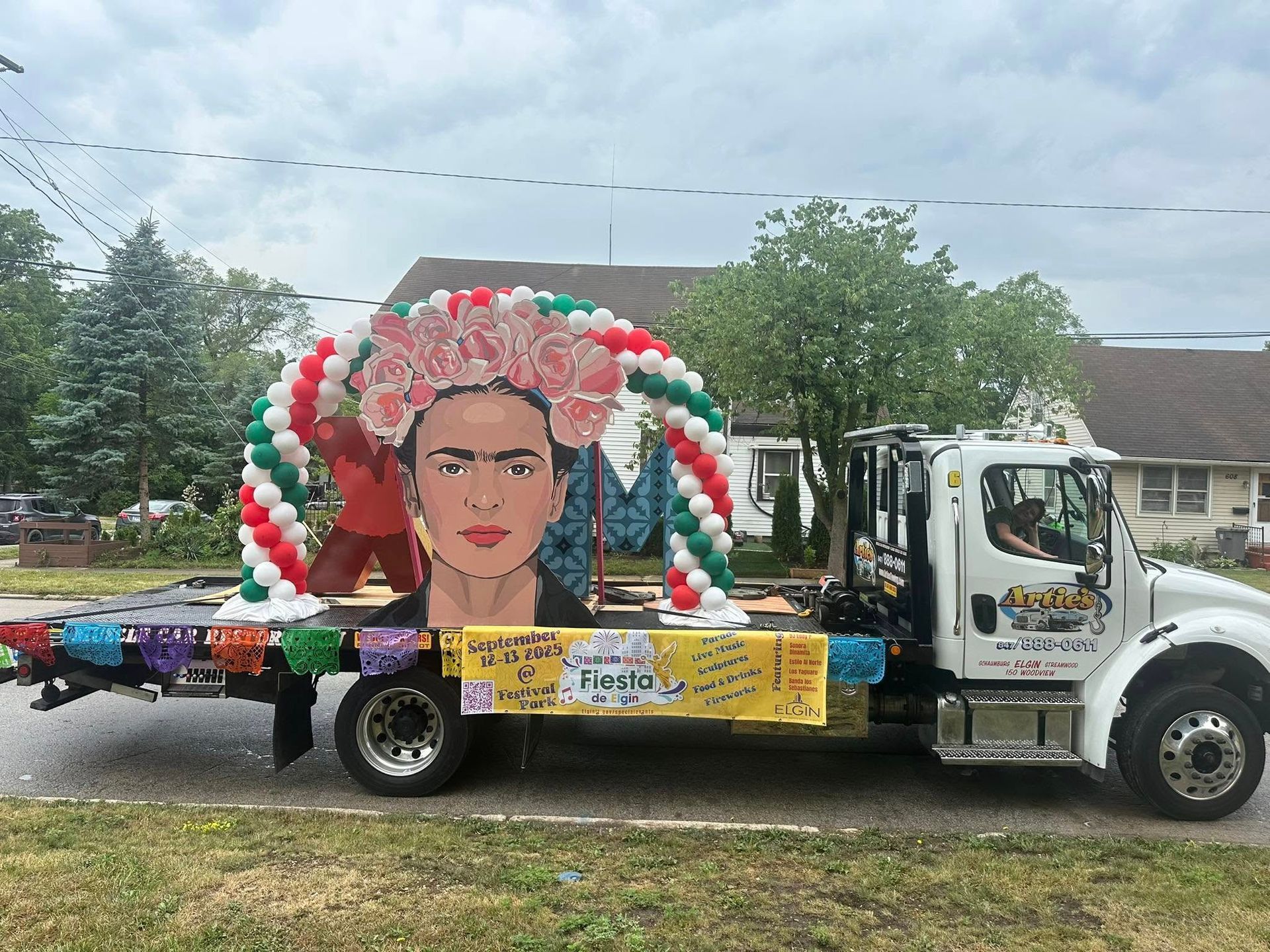 A white tow truck decorated as a Frida Kahlo float with a portrait, balloons, and flowers, driving on a street.