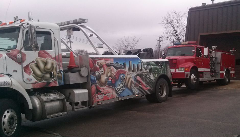 Tow truck towing a red fire truck outside a building on a cloudy day. The tow truck has colorful artwork.