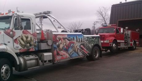 Tow truck towing a red fire truck outside a building on a cloudy day. The tow truck has colorful artwork.