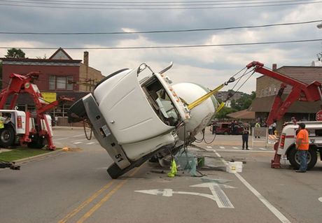 A concrete mixer truck overturned in a street is being uprighted by two tow trucks.