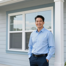 A person in a light blue button-down shirt and dark pants standing on a porch in front of a window.