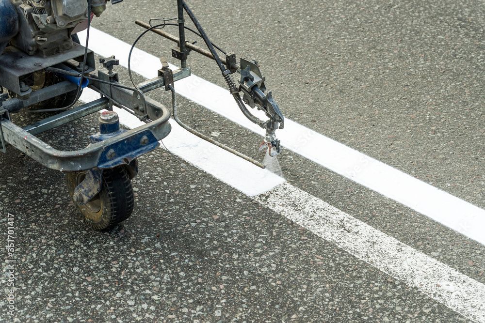 A machine sprays a fresh white line onto an asphalt road next to an existing painted line.