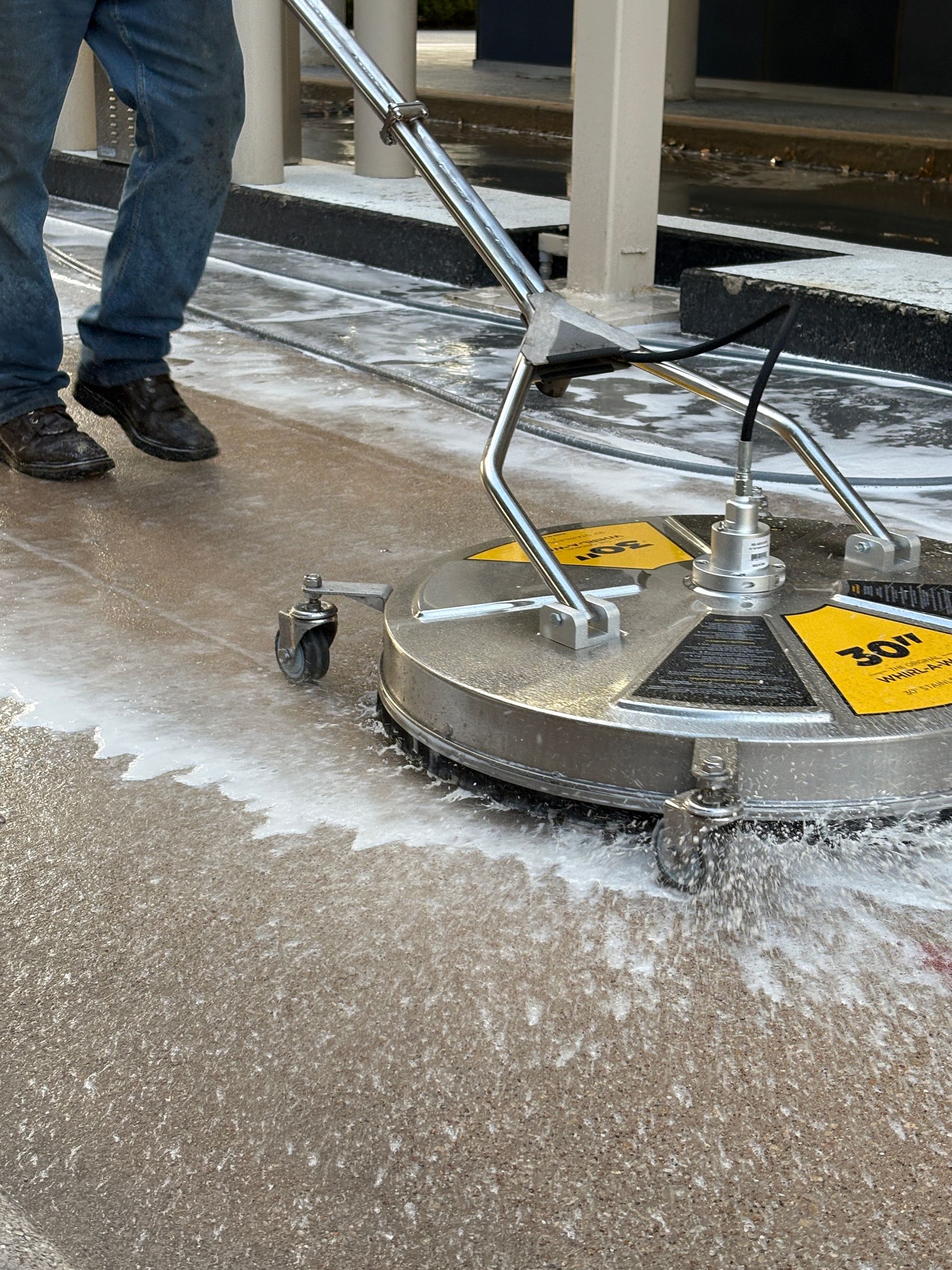 A person uses a circular surface cleaner attachment with a pressure washer to clean a concrete sidewalk.