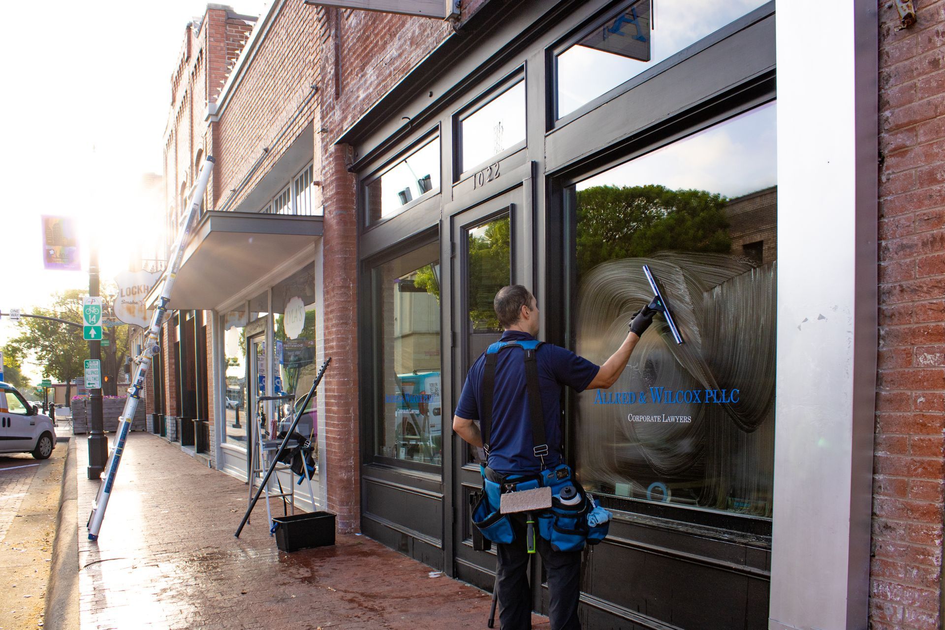 A person in a blue polo shirt uses a squeegee to clean a large storefront window on a sunny street.