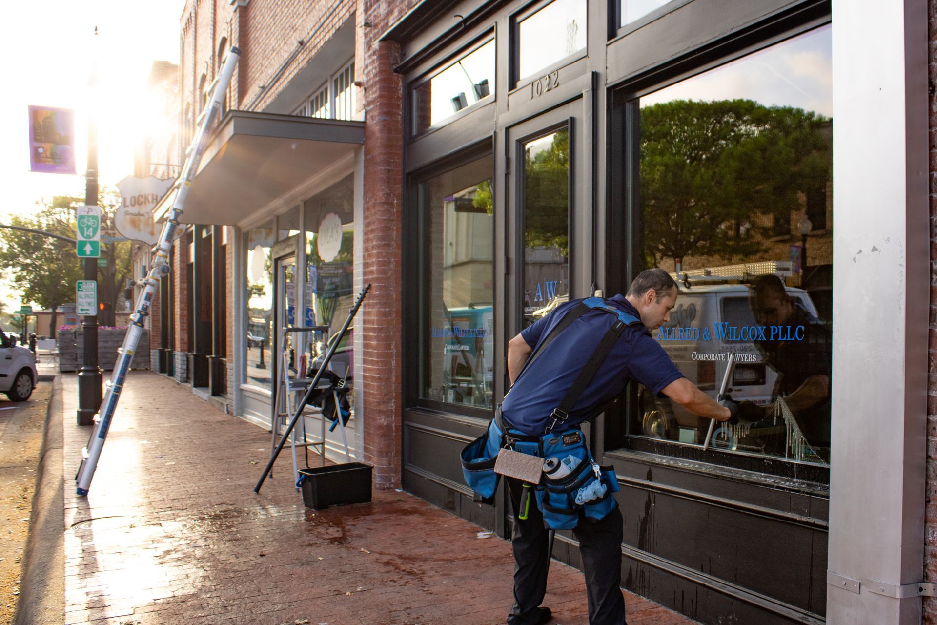 Person in blue works on a storefront window on a sunny brick sidewalk.