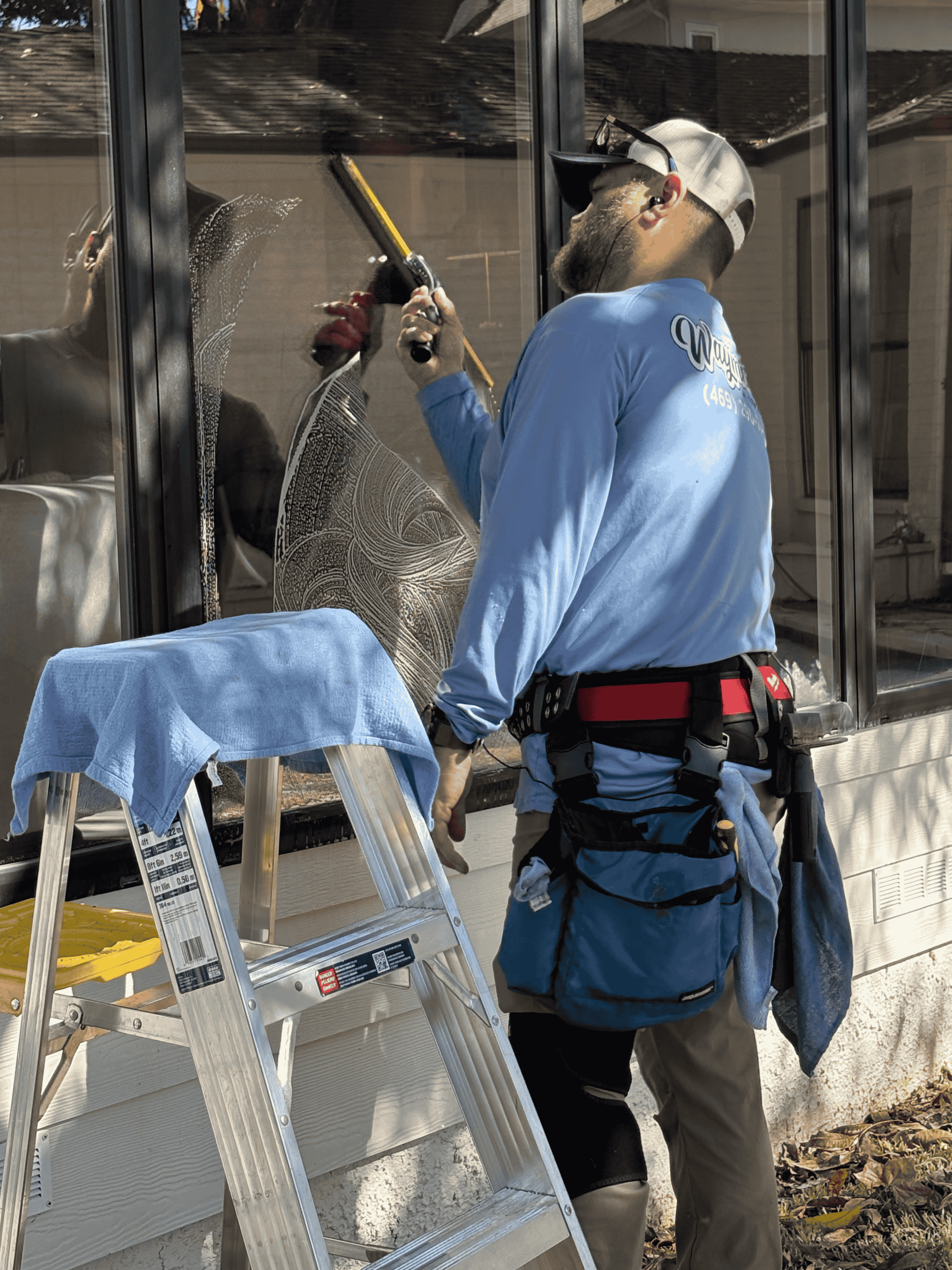 A person in a blue long-sleeve shirt and tool belt cleans a window with a squeegee next to a stepladder.