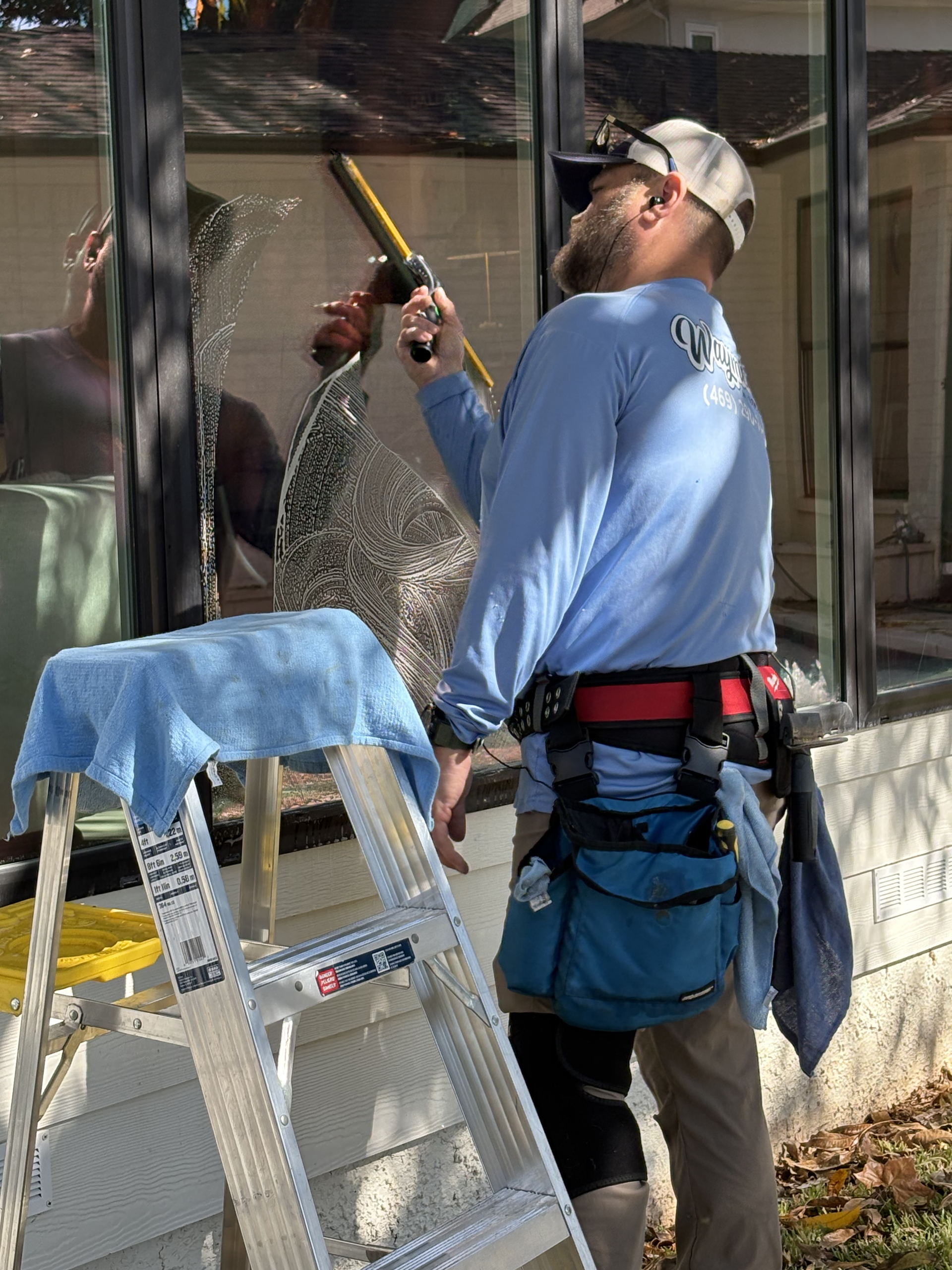 A person wearing a blue shirt and a tool belt uses a squeegee to clean a glass window while standing on a ladder.