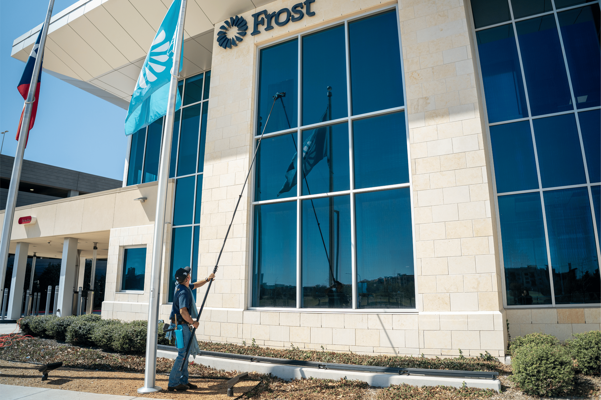 A worker uses a long extension pole to clean the windows of a Frost bank building under a clear blue sky.