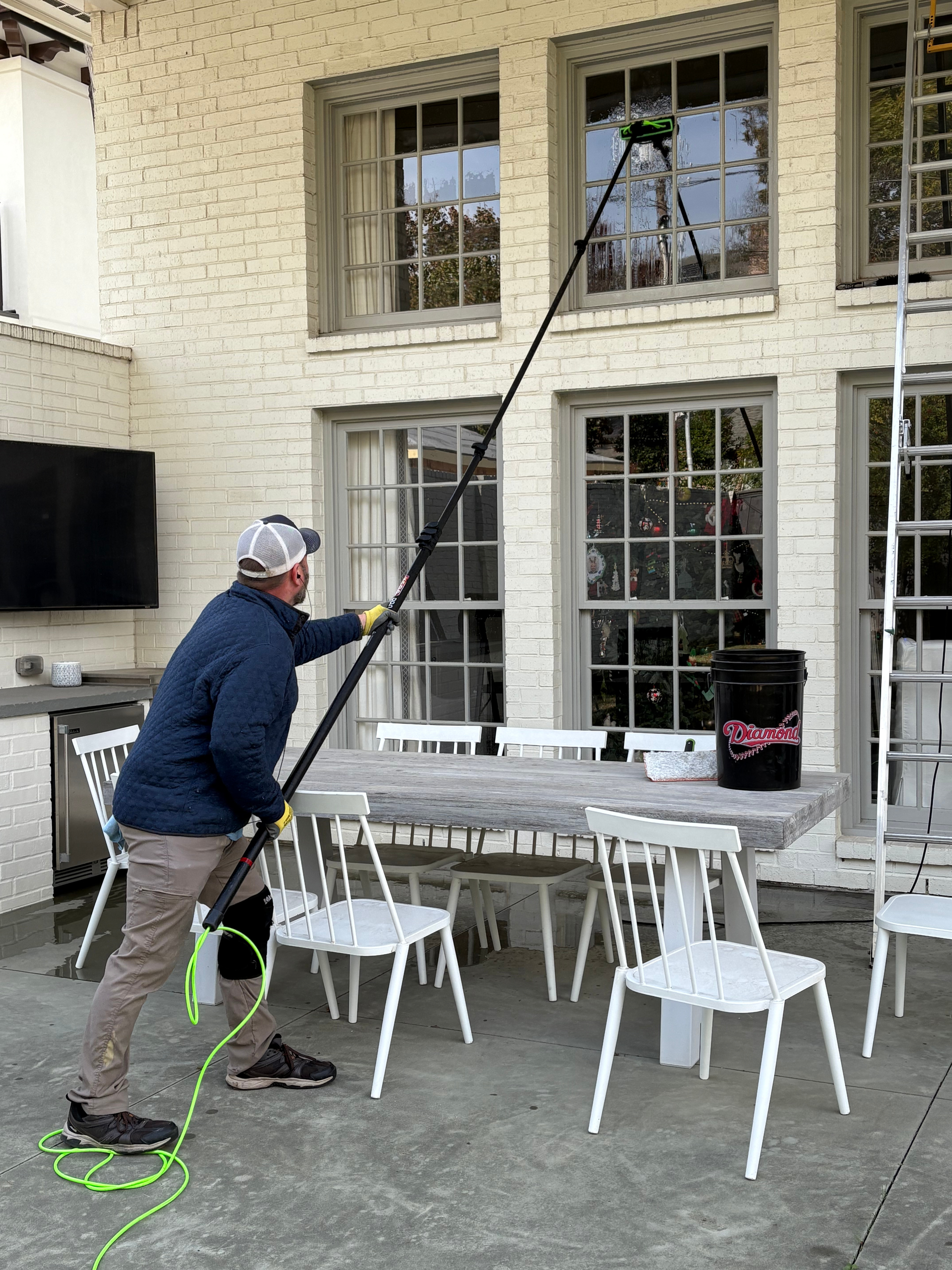 A person cleans a second-story window using a long, telescopic water-fed pole on a patio with a dining set and ladder.