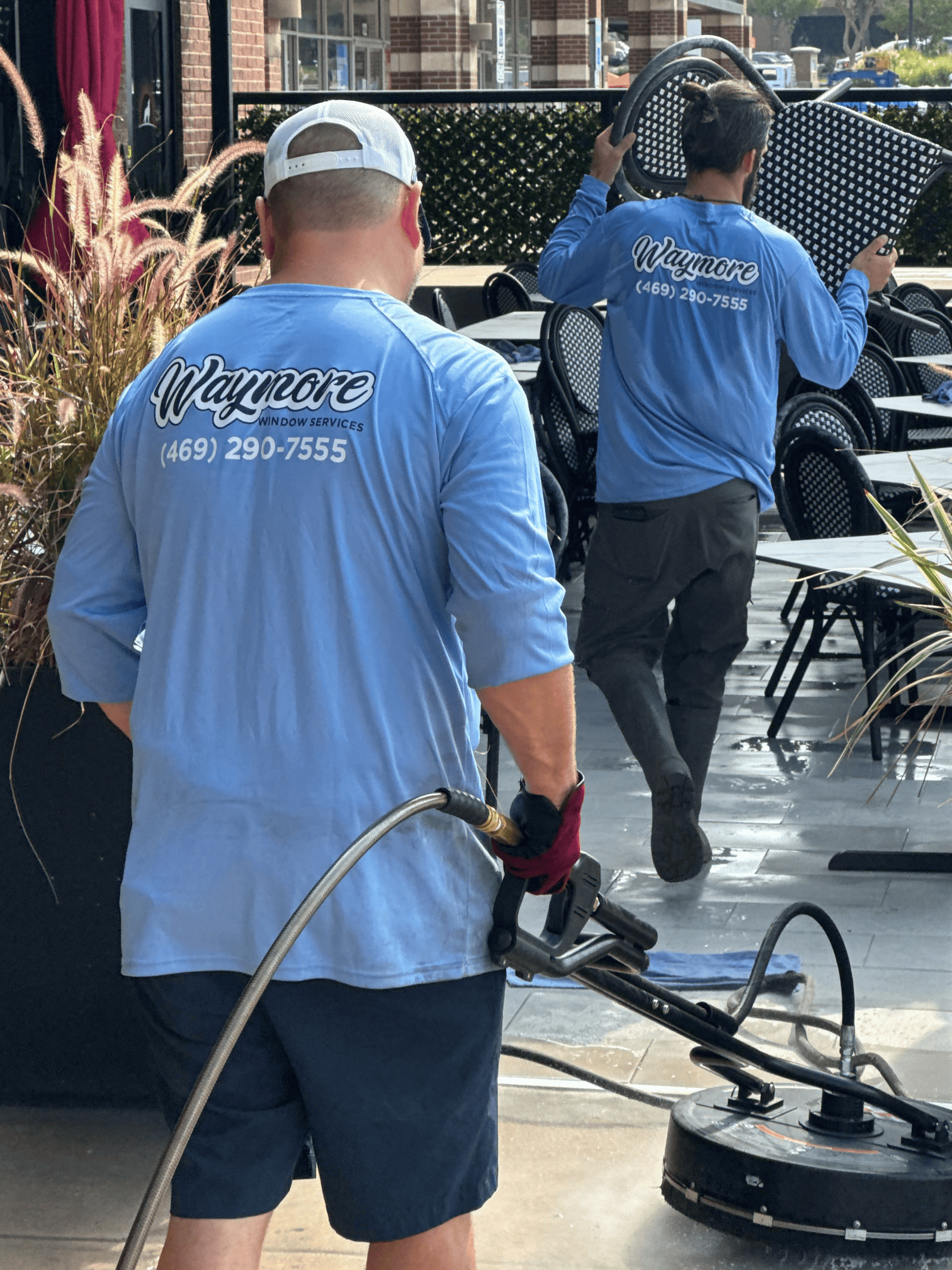Two workers in light blue long-sleeve uniforms pressure wash a restaurant patio floor while moving chairs.